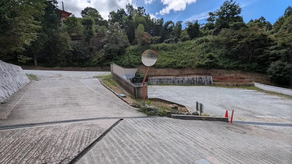 青空の下、緑に囲まれた岡寺の駐車場。急な坂道やカーブミラーが設置され、車でのアクセスの特徴が伺える駐車スペースの様子。