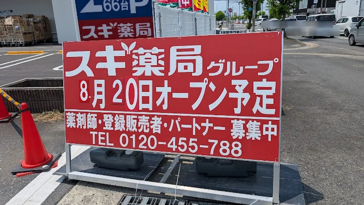 赤い地に白文字で「スギ薬局グループ 8月20日オープン予定」と大きく書かれ、スタッフ募集の電話番号も記載された工事現場の看板のアップ。