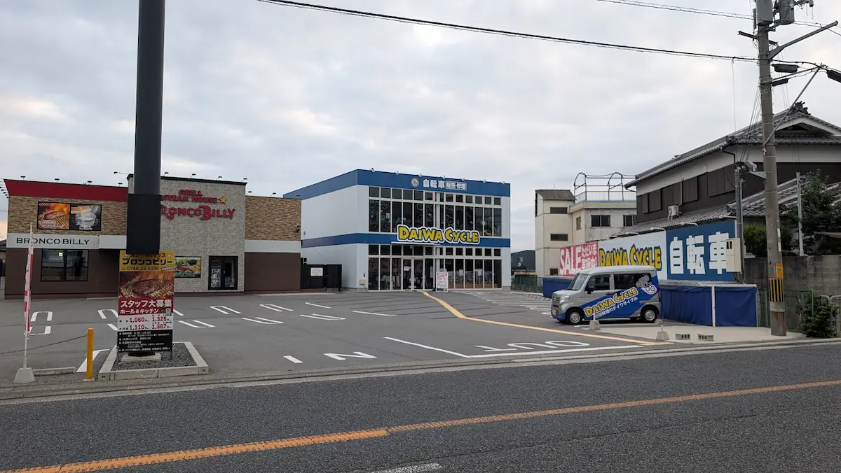 曇天の下、「天下一品」の店舗前から道路を挟んで向かい側に位置する、「ブロンコビリー橿原店」と「ダイワサイクル橿原店」、および手前の広い駐車場の様子。