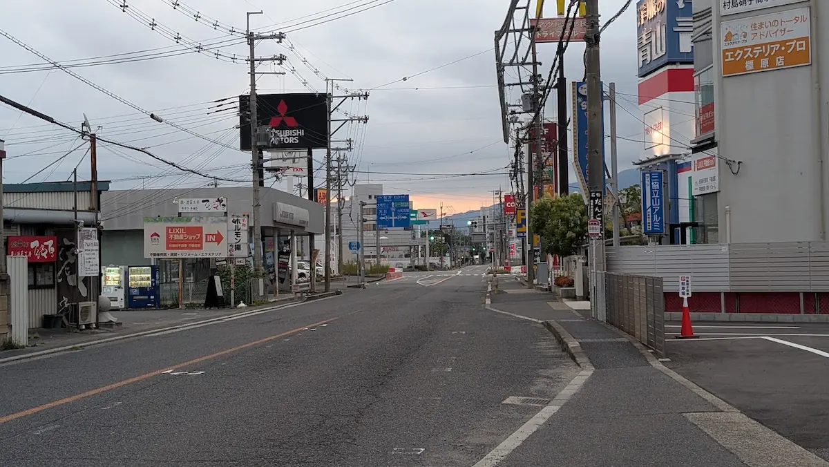 曇天の下、オープン予定の「天下一品」付近から橿原市内方面へ向かう道路の様子。左右にはロードサイドの店舗や看板が立ち並び、遠方に朝日の空が広がる遠景。