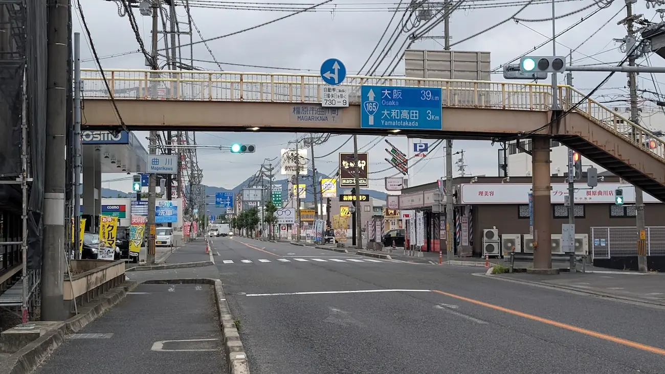 曇天の下、オープン予定の「天下一品」付近から高田方面・大阪方面へ向かう道路の様子。上空には歩道橋や青色の案内標識、左右にはロードサイドの店舗が並ぶ眺望。