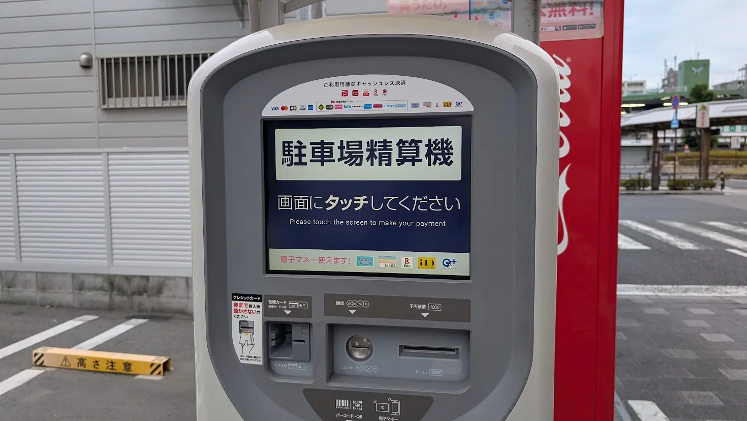 「タイムズ大和八木駅前」の駐車場精算機。画面に「駐車場精算機 画面にタッチしてください」と表示され、各種クレジットカードや電子マネーのロゴが上部や下部に掲示されている様子。