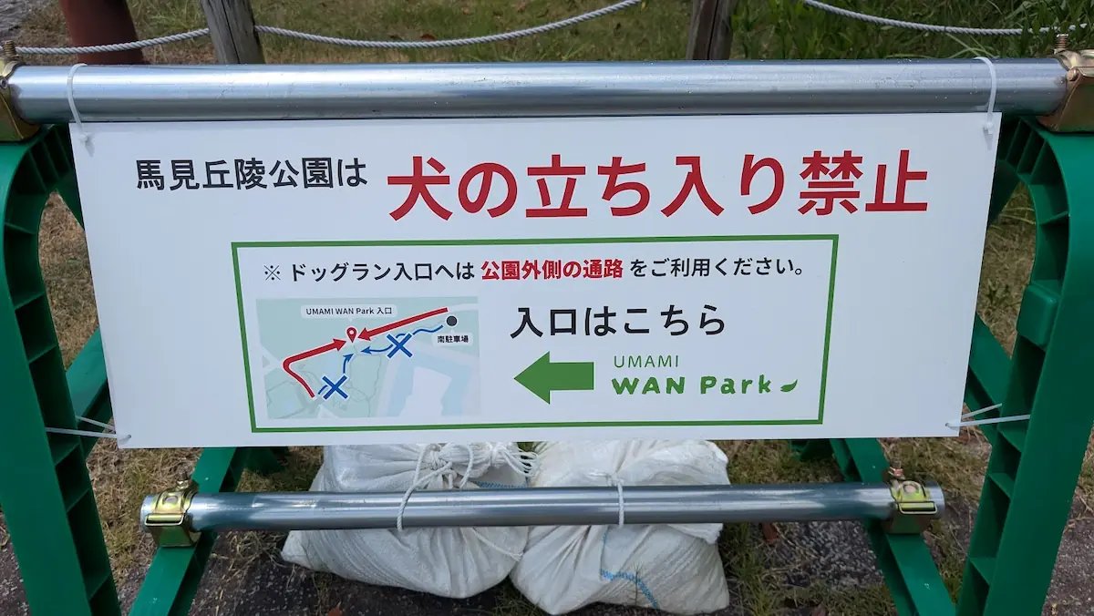 「馬見丘陵公園は犬の立ち入り禁止」と書かれ、新設されたドッグラン「UMAMI WAN Park」へ向かうための公園外側を通る迂回路の案内図が記載された看板。