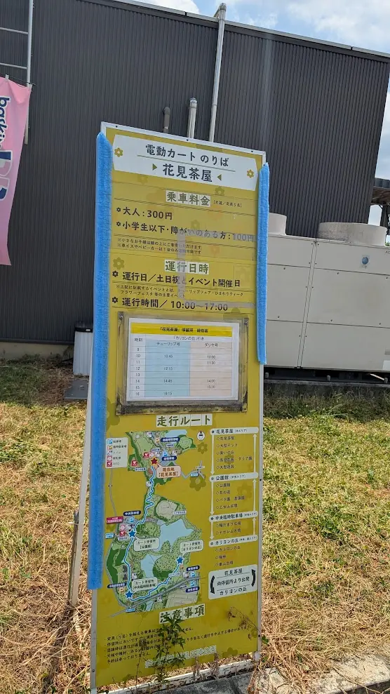 馬見丘陵公園の電動カートのりば（花見茶屋）に設置された、乗車料金、運行日時、時刻表、および走行ルートが記載された黄色の案内看板。