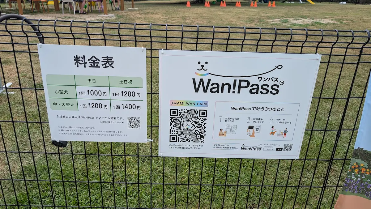 馬見丘陵公園の新設ドッグラン「UMAMI WAN Park」のフェンスに掲示された、平日・土日祝の料金表（小型犬・中大型犬）と、入場・チェックインに必要な「Wan!Pass」のQRコードが記載された案内板の様子。