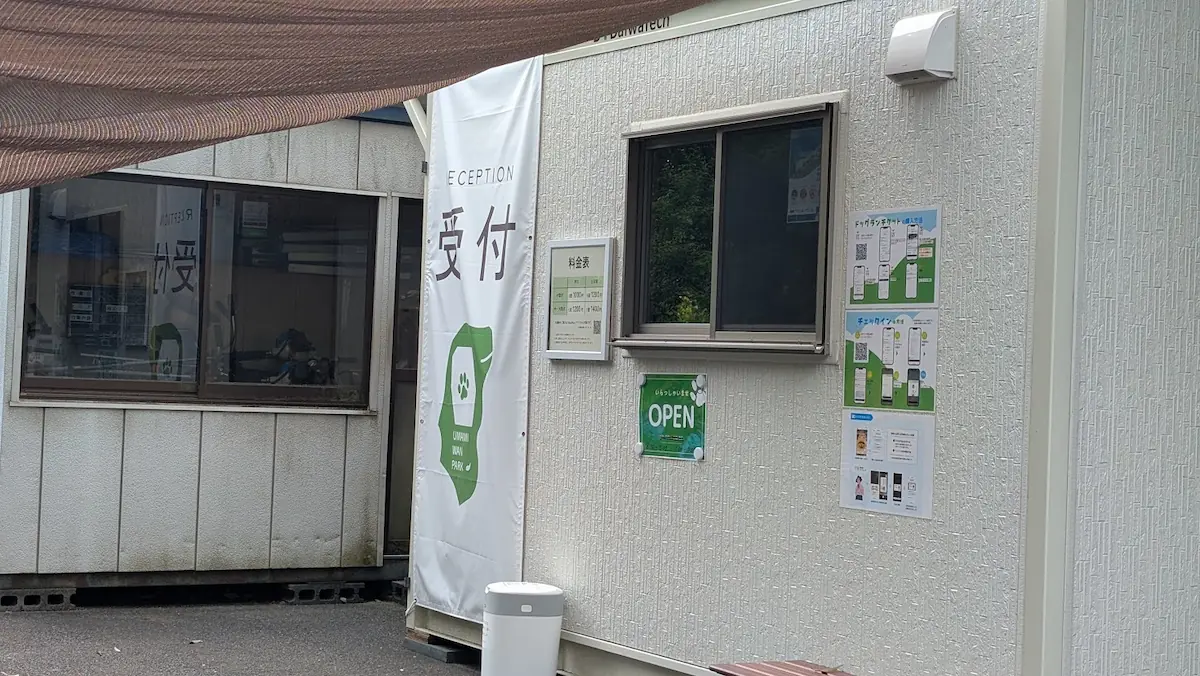 馬見丘陵公園内に新設されたドッグラン「UMAMI WAN Park」の受付建物の外観。大きく「受付」と書かれた白いタペストリーや、料金表、OPENの看板、チケット購入方法の案内が掲示されている様子。