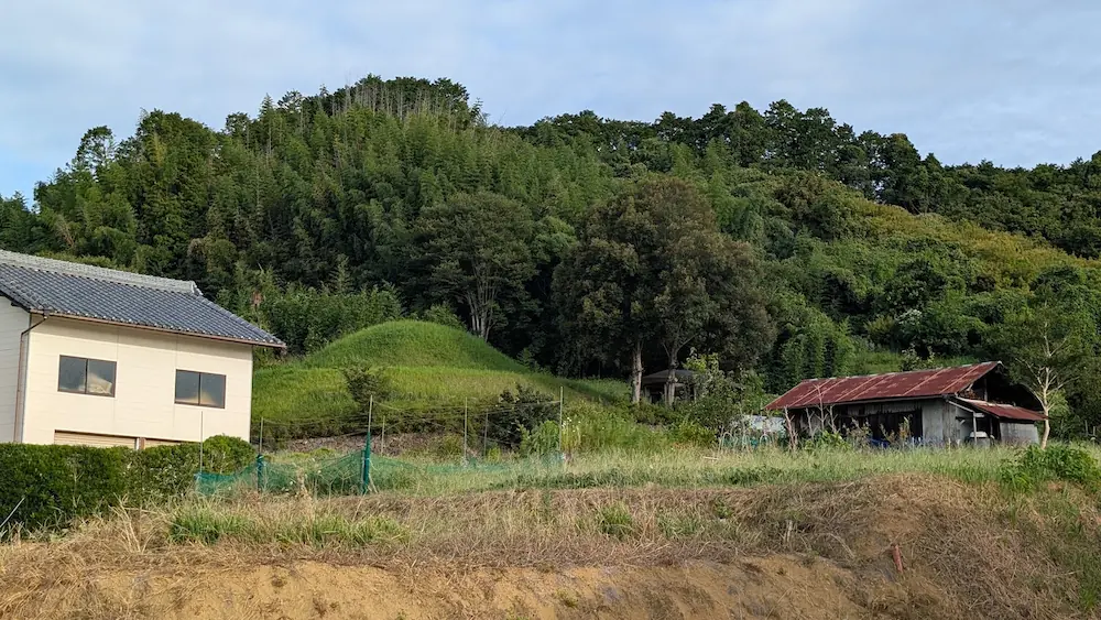 飛鳥村において、民家や畑の背後に緑豊かな山々を背景として佇む「マルコ山古墳」の全景の様子。