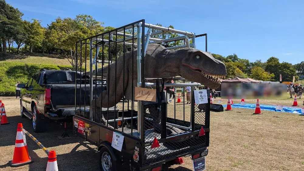 竹取公園のほほえみフェスタ会場において、「ぽち」と書かれた札が掲げられ、檻を積んだトレーラーに載せられて展示されている恐竜のオブジェ。