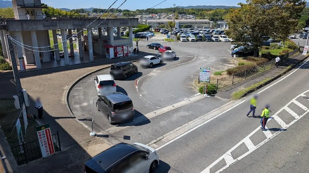 竹取公園のほほえみフェスタ開催日（12時30分）、誘導員が立ち並び駐車場への入庫待ちの車列ができている入り口付近の様子。