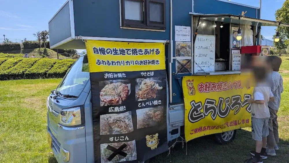 竹取公園のほほえみフェスタ会場において、お好み焼きの大きなバナーが掲げられ、行列ができる人気のキッチンカー「とらじろう」の様子。