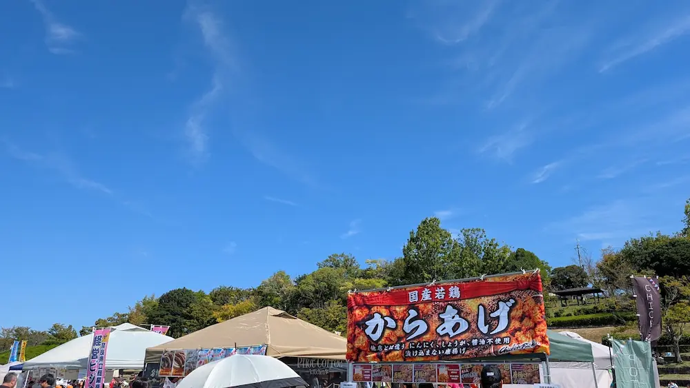 青空の下、竹取公園のほほえみフェスタ会場に並ぶ「国産若鶏からあげ」と書かれた大きな看板が掲げられた屋台・キッチンカーの様子。