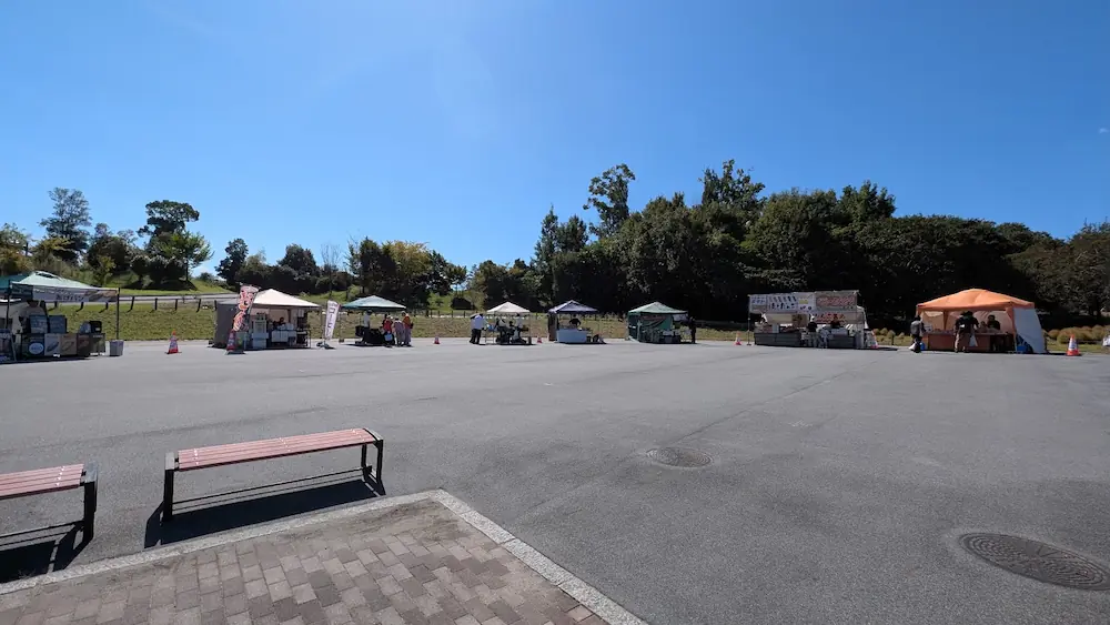 青空の下、馬見丘陵公園の北エリアで開催されている「あきいろマルシェ」において、広場にテントや出店が並んでいる様子。