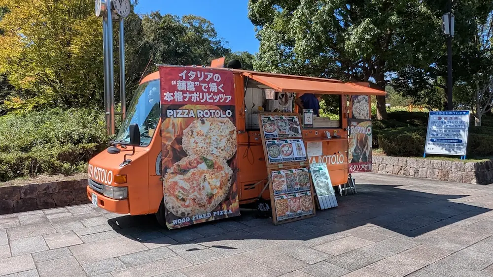 馬見丘陵公園の中央エリアに停車し、「イタリアの薪窯で焼く本格ナポリピッツァ」の看板を掲げたオレンジ色のキッチンカー「PIZZA ROTOLO」の様子。