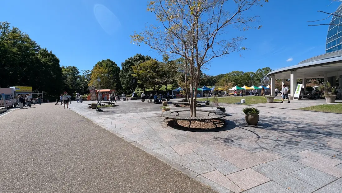 青空の下、馬見丘陵公園の中央エリアの広場において、木製の円形ベンチや多数の出店テントが並んでいる様子。